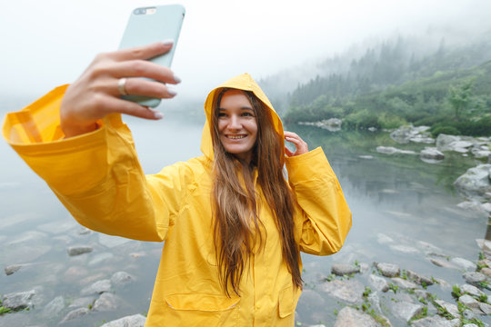 Girl traveler in yellow raincoat with smartphone on a mountain lake. Girl takes photo for travel blog