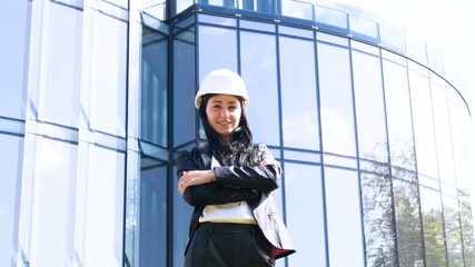 Business woman in suit and helment with crossed arms, looking at camera, smiling. Concept of: Skyscraper background, Communication, Manager, New business.