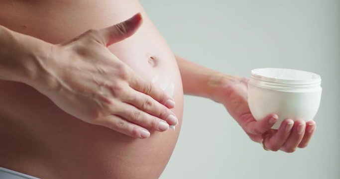 Authentic slow motion close up shot of an young pregnant woman in cosy underwear is applying a moisturizer or sun protection cream on her belly isolated on a white background.