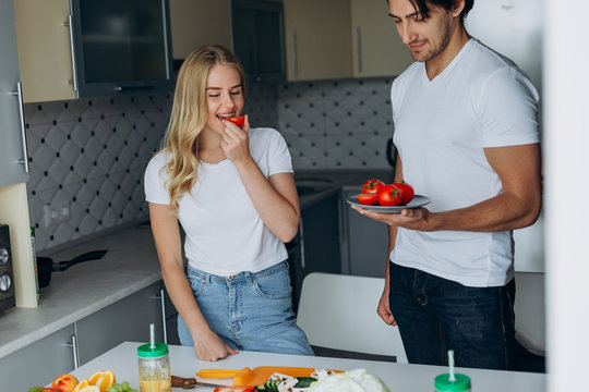 Couple in the kitchen standing  with healthy food. Woman eating a tomato- Image