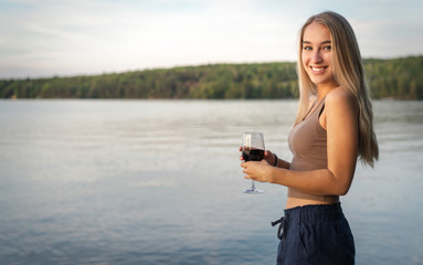 Pretty young woman in summer time with her red glass of wine in front of a calm lake at sunset