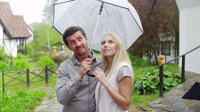 Handheld medium shot of cheerful bearded man and blond woman standing under umbrella and chatting in countryside with lush greenery and rustic houses