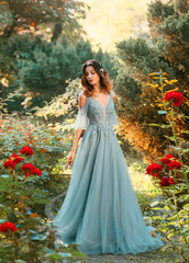 Obraz na płótnie Canvas Affectionate bride in light blue dress in green garden with red lush roses, princess with dark hair looks down, pretty model poses for charming photo with creative colors, symbol of passing summer