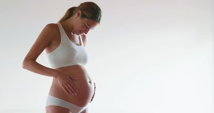 Authentic shot of an young pregnant happy woman in cosy underwear is caressing her belly isolated on a white background.
