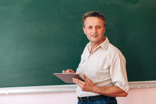 Middle age teacher next the blackboard holding an ipad and explain a lesson.- Image