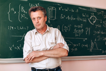 Middle age teacher  standing folded hands next  the blackboard looking at the camera.