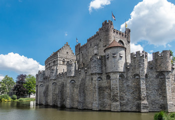 Fototapeta na wymiar Gent, Flanders, Belgium - June 21, 2019: Gray stone cast;e and ramparts of Gravensteen, historic medieval castle of city, behind its moad against blue sky with white clouds. Flags on top, green folia