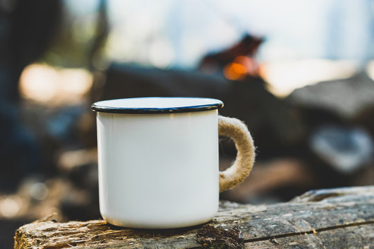 White enamel cup of hot beverage sitting on an old log by an outdoor campfire with a vintage folk edit. Selective focus on mug with blurred background.