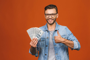 Image of shocked excited young handsome bearded man posing isolated over orange wall background holding money make winner gesture. Thumbs up.