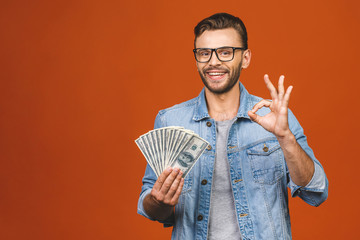 Image of shocked excited young handsome bearded man posing isolated over orange wall background holding money make winner gesture. Ok sign.