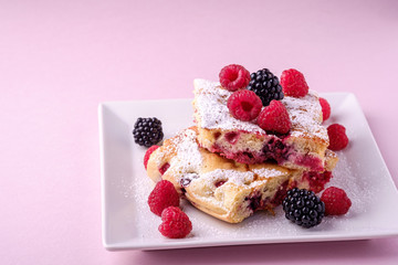 Homemade piece of cake, two pie slices with raspberry and blackberry berries, powdered sugar, in white square plate on pink paper background