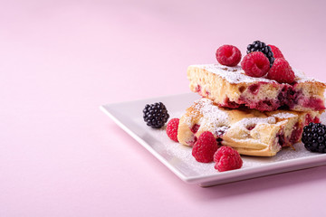 Homemade piece of cake, two pie slices with raspberry and blackberry berries, powdered sugar, in white square plate on pink paper background