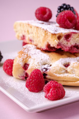 Homemade piece of cake, two pie slices with raspberry and blackberry berries, powdered sugar, in white square plate on pink paper background, macro, close up