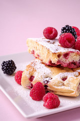 Homemade piece of cake, two pie slices with raspberry and blackberry berries, powdered sugar, in white square plate on pink paper background