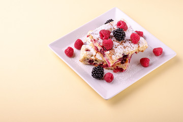 Homemade piece of cake, two pie slices with raspberry and blackberry berries, powdered sugar, in white square plate on cream yellow paper background, copy space