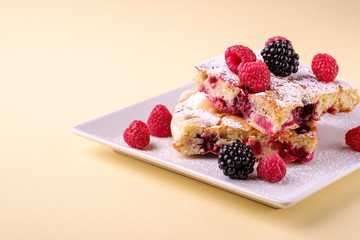 Homemade piece of cake, two pie slices with raspberry and blackberry berries, powdered sugar, in white square plate on cream yellow paper background, copy space
