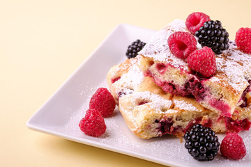 Homemade piece of cake, two pie slices with raspberry and blackberry berries, powdered sugar, in white square plate on cream yellow paper background, copy space