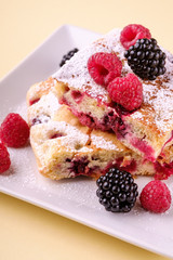Homemade piece of cake, two pie slices with raspberry and blackberry berries, powdered sugar, in white square plate on cream yellow paper background, copy space