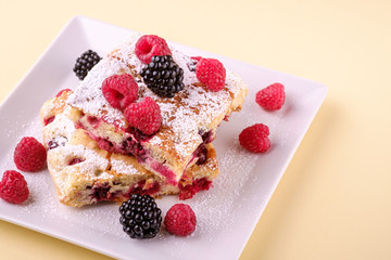 Homemade piece of cake, two pie slices with raspberry and blackberry berries, powdered sugar, in white square plate on cream yellow paper background, copy space