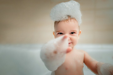 Baby boy bathes in bath with foam and bubbles. Cheerful kid shows his finger to the camera with...