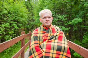 Naklejka na ściany i meble Caucasian male blond stands on a wooden trail in a checkered plaid. Around him are trees, plants. The concept of comfort, unity with the northern nature, Hugge, Lagom.