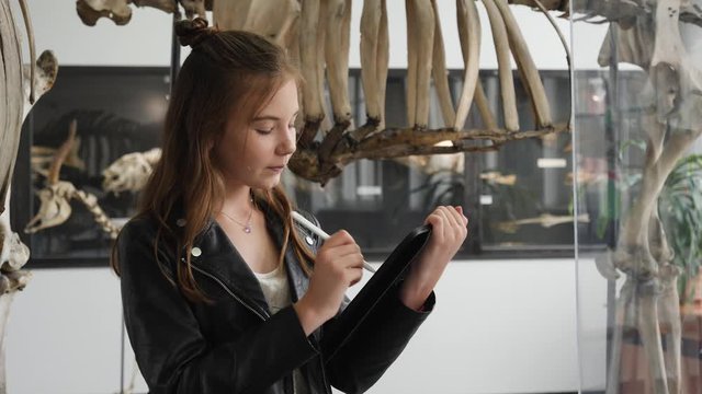 Teenager cute girl makes draw sketches in graphic tablet of animal skeletons in museum of nature as part of school task of biology