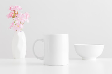 White mug mockup with a bouquet of a pink oleander in a vase and a bowl on a white table.