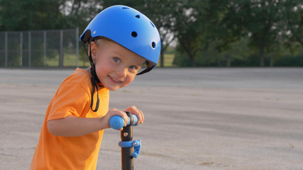 Little boy (kid, child) in orange t-shirt and blue helmet is riding scooter. Childhood memories,...