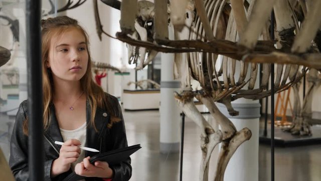 Teenager cute girl makes draw sketches in graphic tablet of animal skeletons in museum of nature as part of school task of biology