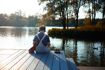 Happy student sitting in front of the city lake in park feel relax and read news on laptop. Boring girl waiting for a boyfriend in autumn romantic day. Quiet island through noise streets. Travel style