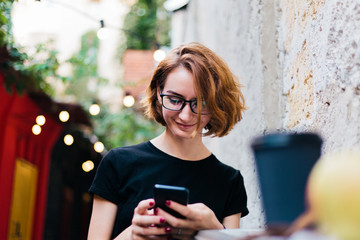 Cheerful hipster girl in glasses with short haircuts use smartphone and sitting in outdoor street...