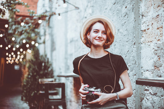 Young hipster female journalist with retro camera  sits on a chair and smiles outdoors against the backdrop of garlands