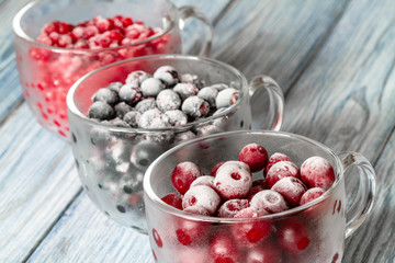 Ripe frozen sweet cherries, red currant and black currant with hoarfrost in the transparent glass cups on blue wooden background. Natural organic healthy food. Closeup, selective focus