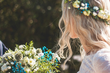 Naklejka premium Beautiful blonde young bride portrait with flower bouquet and wreath on her head in white wedding dress outdoor in summer