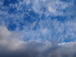 Landscape of beautiful blue sky covered with light thin fluffy white clouds and a thundercloud coming from below. Natural cloudy blue sky on day time abstract background.
