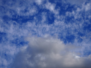 Landscape of beautiful blue sky covered with light thin fluffy white clouds and a thundercloud coming from below. Cloudscape bottom up view. Natural cloudy blue sky on day time abstract background.