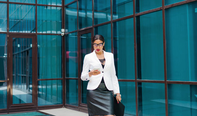 Professional business afro woman in elegant white jacket and skirt with folder with documents in her hands  use smartphone against the background of business center with blue windows