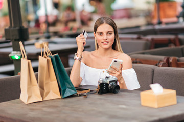 Online shopping. Young shopaholic woman sits at table with shopping bags and holds in her hands smartphone and bank card in outdoor cafe