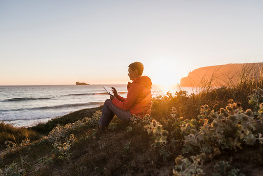 France, Bretagne, Crozon peninsula, woman sitting at the coast at sunset using tablet