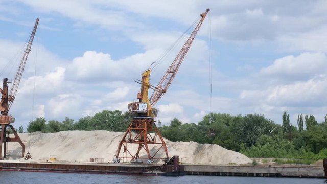 Two port cranes on the river bank produce sand of the river, sunset, barge. Port cranes with a bucket on the river bank, extraction of river sand. Port cargo cranes extracts river sand, production.
