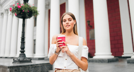 Young fashionable woman holding a cup of coffee on the go against the background of urban architecture with columns