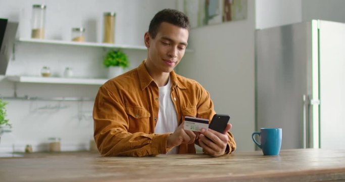 Young Hispanic man is doing online shopping, using credit card and smartphone, happy and smiling, sitting at table in kitchen, latin. 4K, shot on RED camera.