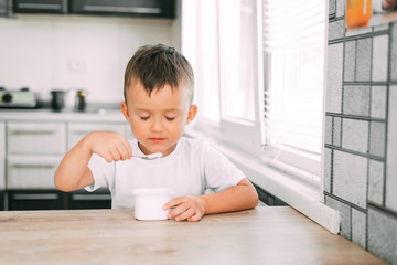 Cute boy in the kitchen eating yogurt from a white yogurt container, a place for advertising