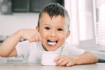 Cute boy in the kitchen eating yogurt from a white yogurt container, a place for advertising