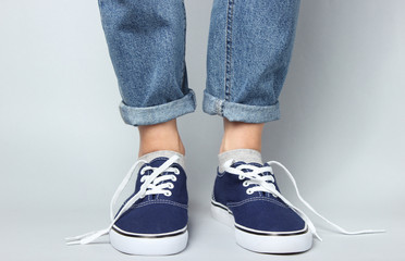 Fashion studio shot. Female legs in jeans and sneakers with untied laces on gray background.