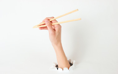 Female hand hold chopsticks through torn hole on white background. Minimalistic food concept. Top view
