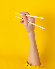 Female hand hold chopsticks through torn hole on yellow background. Minimalistic food concept. Top view
