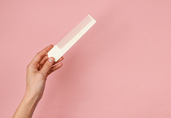 Female hand hold white plastic comb on pink pastel background. Hair care, beauty concept. Top view