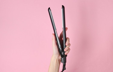 Hair care. Female hand holding hair straightener on pink background. Top view