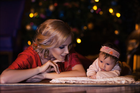 Tender photo of beautiful young mother in red suit with luxurious blond hair posing with her cute little baby girl in studio beside Christmas tree